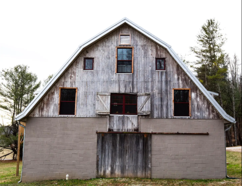 Weathered barn with a wooden upper section and a gray concrete lower section, surrounded by trees.