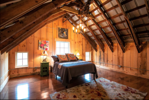 Cozy attic bedroom with wooden walls, a bed, decorative pillows, a rug, and a chandelier. Natural light from windows.
