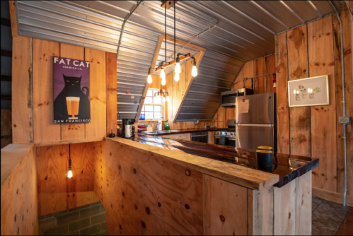 Cozy kitchen with wooden walls, metal roof, and modern appliances, featuring a "Fat Cat" poster and warm lighting.