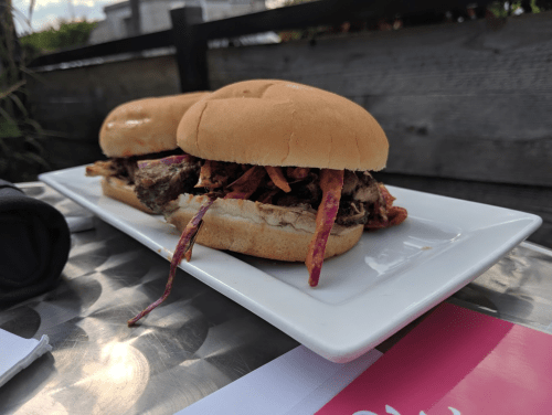 Two pulled pork sandwiches with crispy toppings on a white plate, set on a metal table.