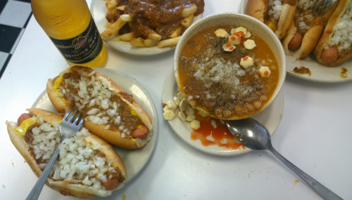 A table with hot dogs topped with onions, chili, fries, and a bowl of chili with toppings, alongside a beer.