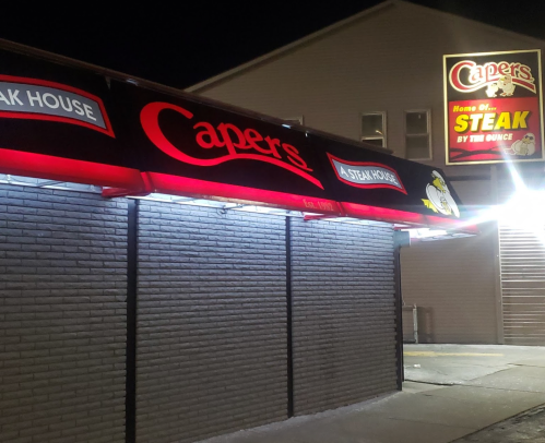A nighttime view of Capers Steak House, featuring a lit sign and awning with the restaurant's name.