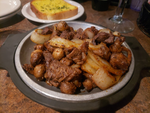 Sizzling platter of beef, onions, and mushrooms, served with a side of garlic bread.