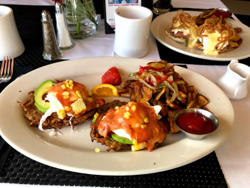 A plate of eggs benedict with corn, avocado, and a side of crispy potatoes, garnished with a strawberry and orange slice.