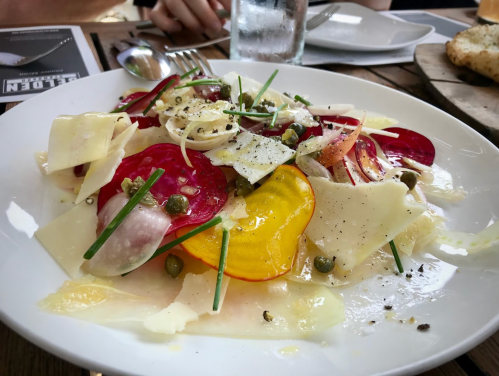 A colorful salad with sliced beets, cheese, capers, and herbs on a white plate, served on a wooden table.