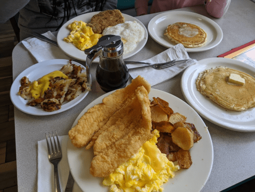 A table with plates of breakfast food: eggs, pancakes, hash browns, bacon, and fried fish, with syrup in a glass jar.