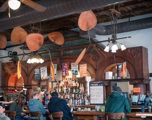 A cozy bar interior with patrons sitting at the counter, surrounded by wooden decor and a variety of drinks on display.