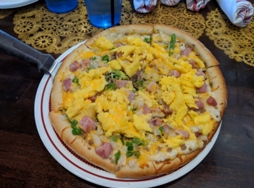 A pizza topped with scrambled eggs, ham, green onions, and melted cheese on a golden crust, served on a plate.
