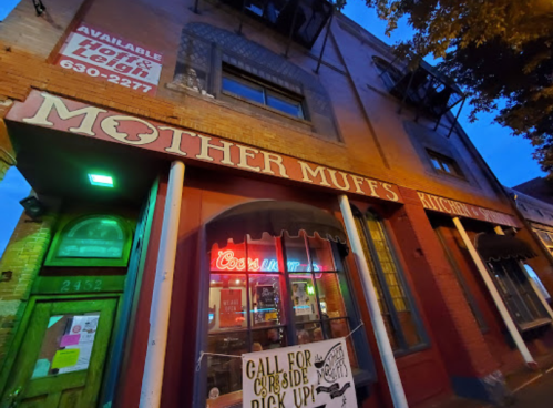 Exterior of Mother Muff's Kitchen at dusk, featuring a neon sign and a sign for curbside pickup.
