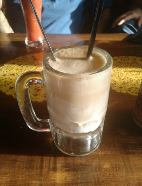 A frothy beverage in a glass mug with two straws, sitting on a wooden table with a lace doily.