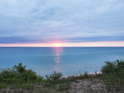 A serene beach scene at sunset, with calm waters reflecting the colorful sky and lush greenery in the foreground.