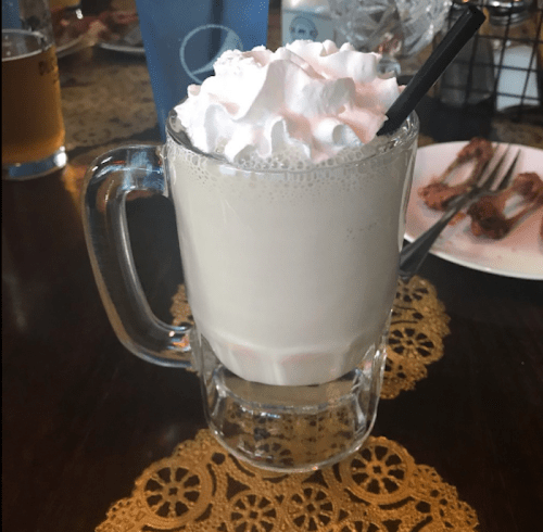 A frosty glass mug filled with a creamy beverage topped with whipped cream and a straw, set on a table.