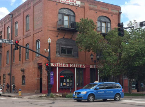A red brick building with a sign for "Other Muffs" and a blue taxi at a traffic light on a city street.