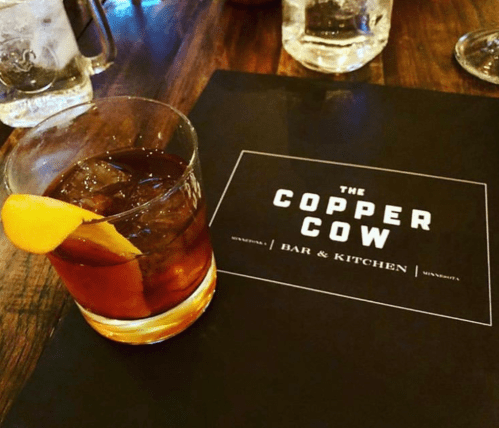 A cocktail with an orange slice on a wooden table next to a menu for "The Copper Cow Bar & Kitchen."