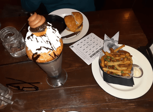A dessert with whipped cream and a cookie on top, alongside a burger and a basket of fries on a wooden table.