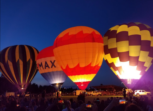 Three colorful hot air balloons illuminated at dusk, with a crowd gathered below and a clear blue sky above.