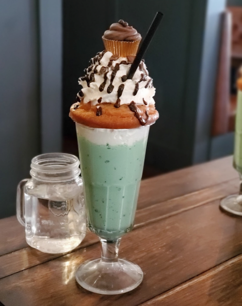 A tall glass filled with mint green milkshake, topped with whipped cream, chocolate drizzle, and a mini cupcake.
