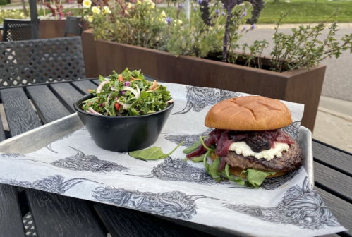 A gourmet burger with greens and a side salad on a metal tray, set on a table with plants in the background.