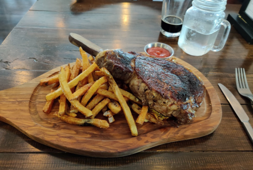 A large steak on a wooden platter with crispy fries, a small bowl of ketchup, and drinks in the background.