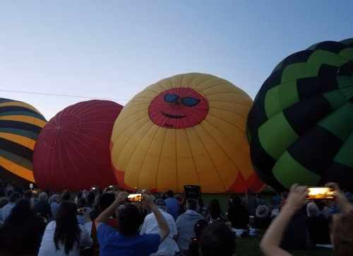 A crowd watches colorful hot air balloons, including a smiling yellow one, at dusk.