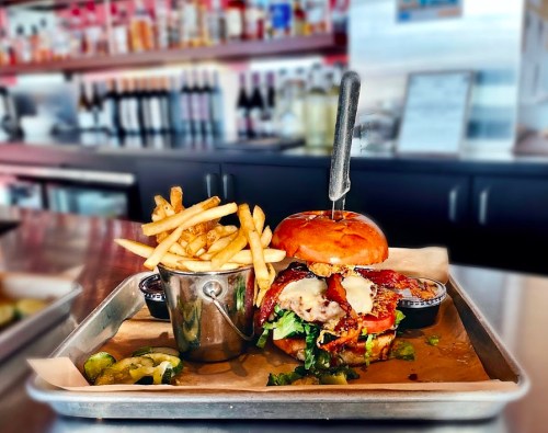 A gourmet burger with bacon and cheese, served with fries in a metal cup, on a tray in a restaurant setting.