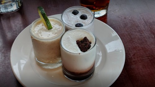 Three small dessert cups on a plate: one with a lime garnish, one with blueberries, and one with chocolate crumbs.