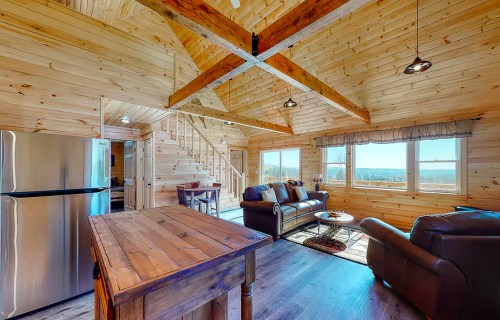 Cozy wooden interior of a cabin with a kitchen, living area, and large windows showcasing a scenic view.