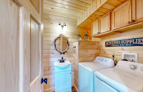 A cozy laundry room with wooden walls, a round mirror, a light blue sink, and a washing machine.