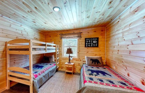 Cozy wooden bedroom with bunk beds and a single bed, decorated with bear-themed quilts and a lamp by the window.