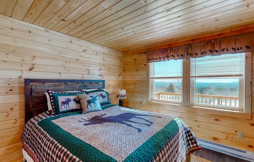 Cozy bedroom with wooden walls, a patterned quilt featuring moose, and large windows offering a scenic view.