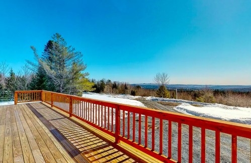 A wooden deck with a red railing overlooks a snowy landscape and clear blue sky.