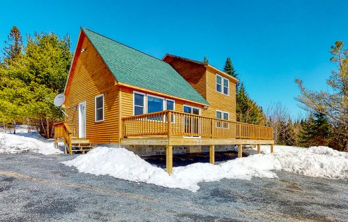 A wooden cabin with a green roof, surrounded by snow and trees, featuring a large deck and clear blue sky.
