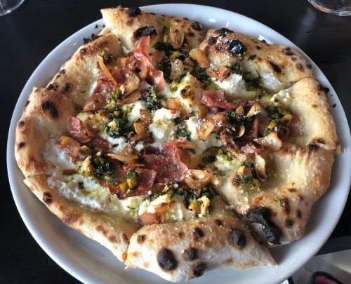 A freshly baked pizza topped with cheese, prosciutto, garlic, and herbs on a golden crust, served on a white plate.