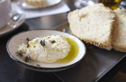 A plate with creamy cheese drizzled with olive oil, garnished with herbs, alongside slices of fresh bread.
