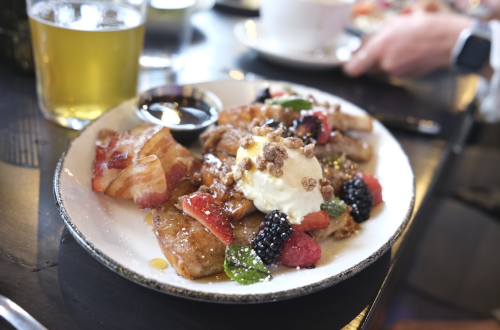 A plate of French toast topped with berries, whipped cream, and granola, served with bacon and syrup on the side.