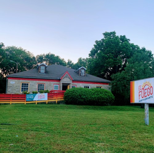 A stone building with red trim, featuring a sign for "El Fuego Restaurante" and a grassy area in front.