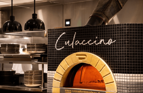 A pizza oven with a tiled exterior, featuring the word "Culaccino" and surrounded by kitchen equipment and plates.