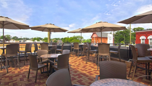 A rooftop patio with tables and umbrellas, featuring a brick floor and a view of nearby buildings and trees.
