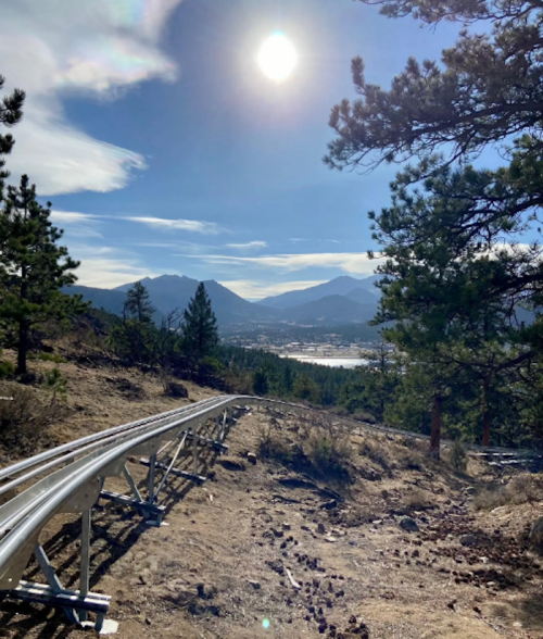 A scenic view of a mountain landscape with a metal track winding through trees, under a bright sun and blue sky.