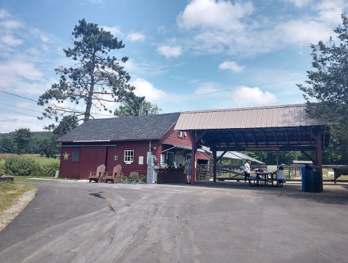 A red barn-style building with a covered area, surrounded by trees and a clear blue sky. A person is seated at a table.