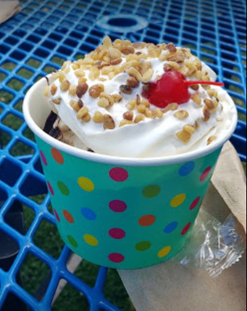 A colorful cup of ice cream topped with whipped cream, chopped nuts, and a cherry, sitting on a blue table.