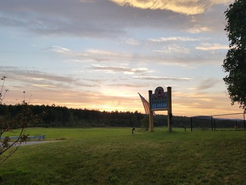 A sunset over a grassy field with a sign for an ice cream shop in the foreground.