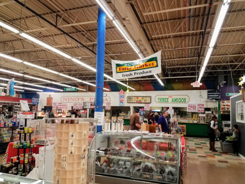 A bustling market interior with various food stalls, colorful signage, and shoppers browsing fresh produce and other goods.