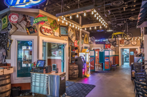 A vibrant bar interior featuring colorful signs, a gaming area, and rustic decor with barrels and string lights.