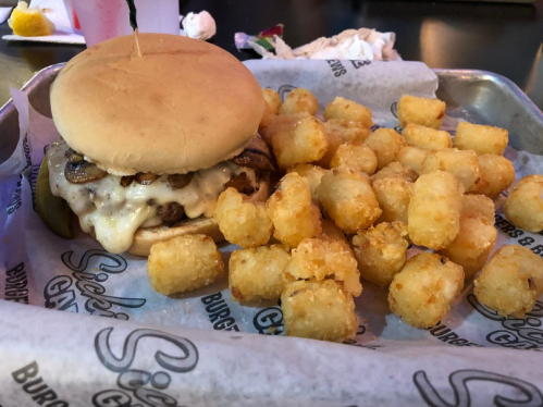 A cheeseburger with mushrooms and pickles served with a side of crispy tater tots on a tray.