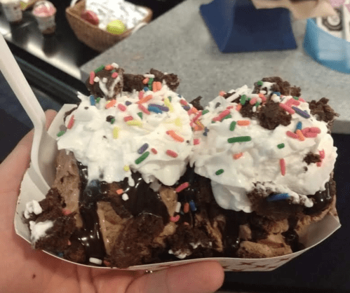 A hand holding a dessert bowl filled with chocolate ice cream, whipped cream, chocolate sauce, and colorful sprinkles.