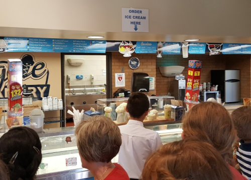 A busy ice cream shop with a counter, menu overhead, and customers waiting in line to order.
