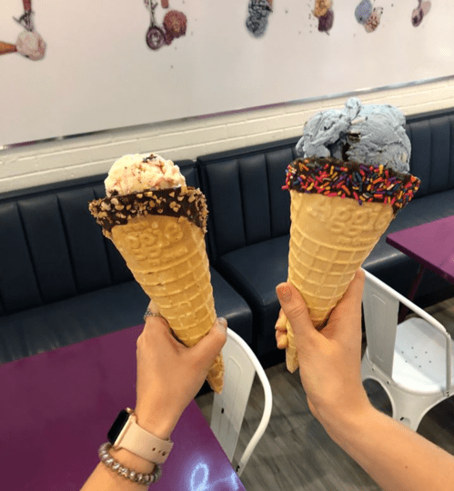 Two hands hold large ice cream cones, one with chocolate-dipped and sprinkles, the other with blue and red flavors.