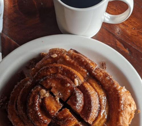A cinnamon roll drizzled with icing on a plate, next to a steaming cup of coffee.