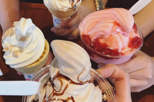 Four hands holding different ice cream cups: soft serve, strawberry swirl, and a cone topped with nuts.